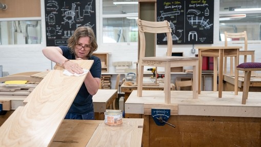 A student sanding a large plank of wood with furniture on display in the background.