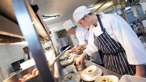 Students in chef's whites and aprons plating food in a kitchen.