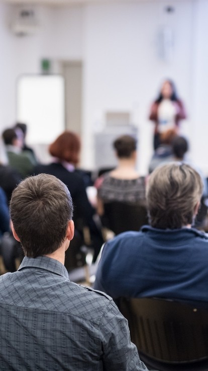 Ystafell o bobl yn eistedd gan wynebu ymlaen tuag at siaradwr yn ystod seminar