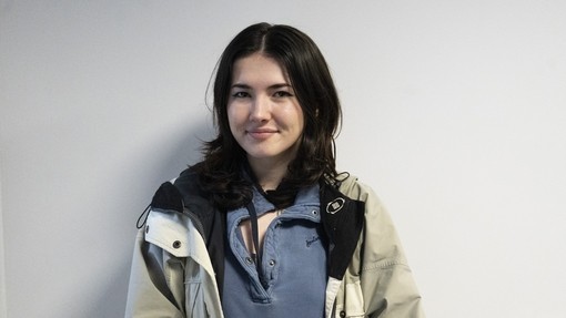 Olivia standing against a front wall looking at the camera in a blue top and coat