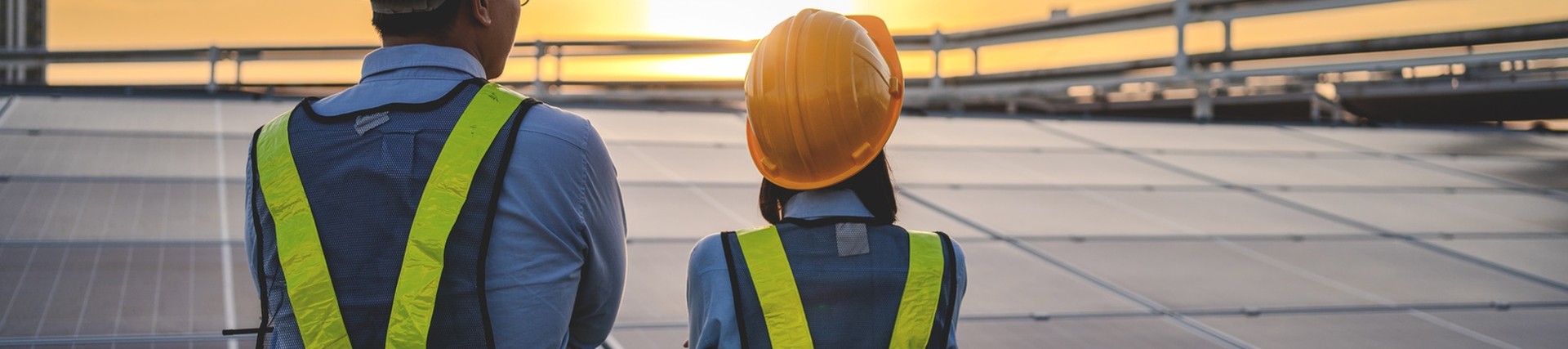 Two managers, one male, one female. Both wearing hivis and har hats while looking towards solar panels, their back turned to the camera