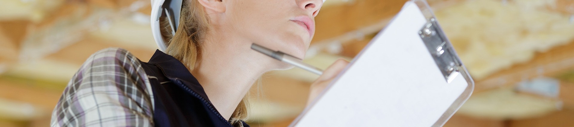 Female worker wearing a white hat writing on a clipboard while looking at the wooden ceiling of a construction site