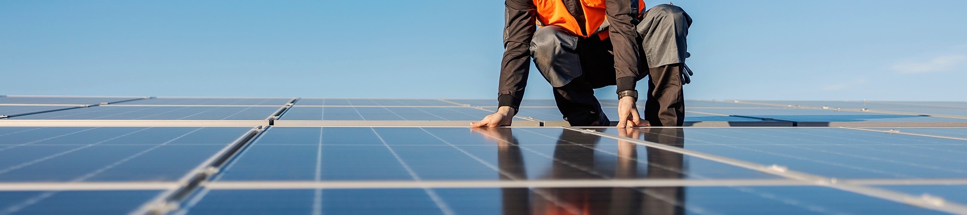 Male installer wearing orange hi vi and white hard hat inspecting solar panels on a clear blue day