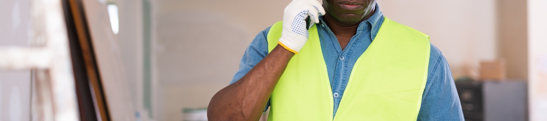 Black retrofit assesor on a mobile phone in a construction site. He's wearing a white hard hat and hivis bib